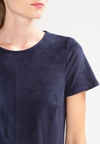 Haut bleu marine à manches courtes en tissu semblable au daim. Présente une texture lisse et des détails discrets de coutures sur la poitrine.