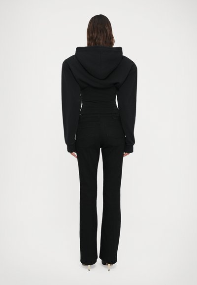 Donna che indossa un cropped hoodie nero e jeans neri a vita alta, in piedi con le spalle rivolte verso la fotocamera su uno sfondo bianco semplice.