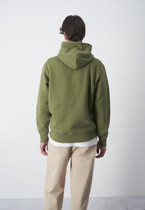 Uomo che indossa una felpa con cappuccio verde oliva, pantaloni beige e una maglietta bianca, in piedi di spalle su uno sfondo chiaro e semplice.
