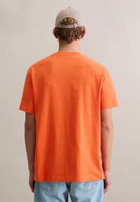 Pomarańczowy bawełniany t-shirt z okrągłym dekoltem i krótkimi rękawami, pokazany od tyłu. W połączeniu z jasnymi jeansami. Gładka tekstura.