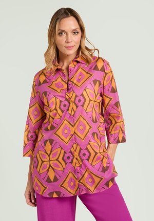 Donna che indossa una camicia a maniche lunghe con grandi motivi floreali geometrici arancioni e rosa e pantaloni rosa acceso su uno sfondo semplice.
