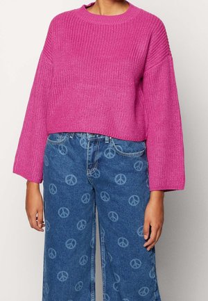Roze gebreide trui met een ronde hals en ruime mouwen, gecombineerd met blauwe jeans van denim met een vredessymboolpatroon.