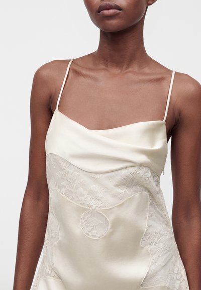 Femme portant une robe en satin crème avec des détails en dentelle et de fines bretelles spaghetti, vue du cou jusqu'au milieu du torse sur un fond uni.