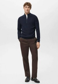 Marineblauer, gerippter Pullover mit Reißverschluss bis zur Brust, weißes Innenfutter, braune gerade geschnittene Hose und schwarze Lederschuhe vor einfachem Hintergrund.