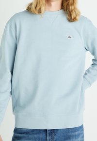 Sudadera de color azul claro hecha de una mezcla de algodón, con cuello y puños acanalados, y pequeño logo bordado "Lee" en el pecho.