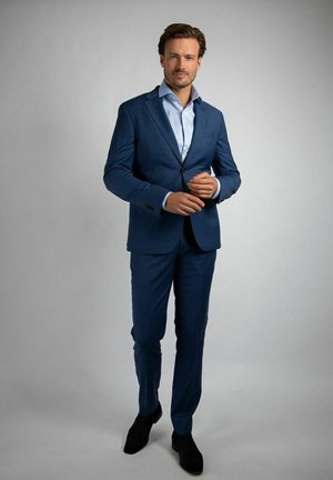 Man die een blauw pak en lichtblauw overhemd draagt, staat tegen een effen grijze achtergrond en past zijn jasje aan bij de mouw.