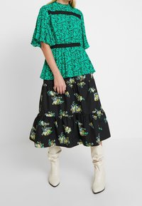 Blusa verde con patrón y mangas acampanadas, combinada con una falda negra de flores con volantes y botas crema hasta la rodilla. Acentos de encaje negro en la blusa.