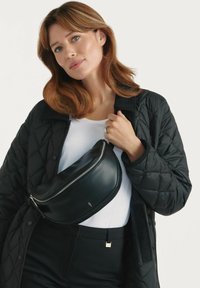 Czarna skórzana torba na ramię o zakrzywionym kształcie, z zapięciem na zamek i małym logo. Nosi się ją na białej koszuli i czarnej pikowanej kurtce.