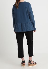 Top blu con maniche lunghe, dettagli in occhio di pernice e orlo a peplum, abbinato a pantaloni neri cropped e sandali platform color camoscio. Tessuto di una consistenza liscia.