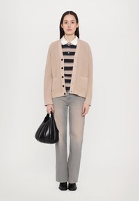 Μπεζ πλεκτό κα cardigan με μπροστινές τσέπες, συνδυασμένο με μαύρο και λευκό ριγέ πουκάμισο. Το μοντέλο κρατά μια μαύρη πτυχωτή τσάντα, φορώντας ανοιχτόχρωμα τζιν.