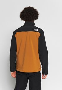 Veste en polaire avec une partie supérieure noire et une section inférieure orange. Col montant, fermeture zippée et logo The North Face au dos.
