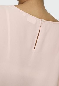 Jasnoróżowa bluzka z dekoltem w kształcie łezki na plecach z pojedynczym zapięciem na guzik, wykonana z finezyjnego, teksturowanego materiału o miękkim układzie.