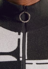 Gros plan sur la fermeture éclair d'un pull en maille noir et blanc avec un anneau de tirage circulaire gravé du mot HUGO.