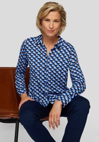 Hemd mit Knöpfen in blau-weißem geometrischem Muster, mit langen Ärmeln, einem Kragen und einer taillierten Silhouette. Kombiniert mit dunklen Hosen.