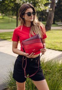 Červená a vínová taška přes tělo s horizontálními pruhy, vyrobena z hladké kůže, se stříbrnými detaily a nastavitelným popruhem. Logo je viditelně umístěné.