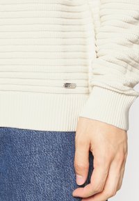 Gros plan d'une main reposant à la hanche portant un pull côtelé crème avec une petite étiquette métallique « HUGO » et un jean en denim bleu.