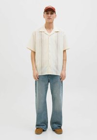 Camisa de botones de manga corta color crema con rayas verticales, combinada con jeans de mezclilla azul de pierna ancha y zapatos marrones sin cordones.