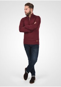 Burgundska dukserica na zip s dugim rukavima, rebrastim rubom i kožnom oznakom. Nosi se uz tamnoplave traperice i smeđe zamršene gležnjače.