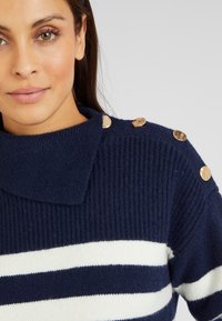 Maglione a coste blu navy con strisce bianche, colletto pieghevole e bottoni decorativi dorati sulla spalla. Texture morbida e design casual.