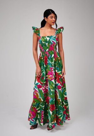 Mujer con vestido maxi sin mangas de colores vibrantes, con tirantes con volantes y un patrón de hojas tropicales en verde, rosa y rojo.