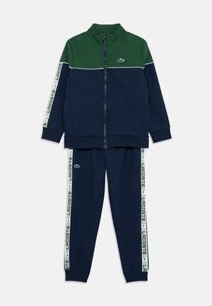 Ensemble de survêtement bleu marine et vert avec veste zippée et pantalon. Doté d'une bande blanche avec le logo "LACOSTE" et d'un logo brodé sur la poitrine. Texture douce.