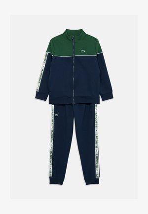 Navy a zelený sportovní oblek s krátkým zipem, zahrnující bundu a kalhoty. Vybavený bílým pruhem s logem "LACOSTE" a vyšívaným logem na hrudi. Jemná textura.