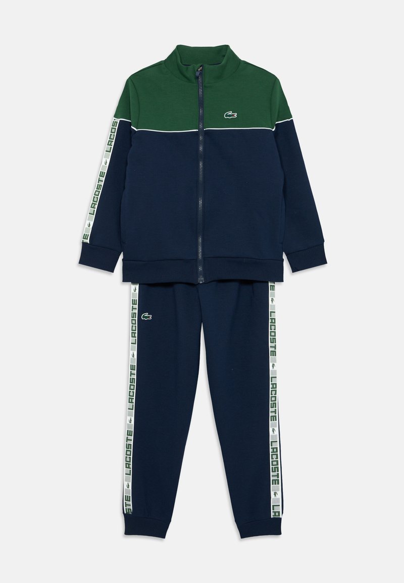 Training suit albastru închis și verde, cu jachetă cu fermoar și pantaloni. Prezintă o dantură albă cu logo-ul "LACOSTE" și logo brodat pe piept. Textură moale.