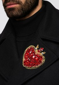 Rote brosche in Herzform mit Kristallen, goldenen Akzenten und Kronen-Design, an einem schwarzen Mantel über einem schwarzen Rollkragenpullover befestigt.