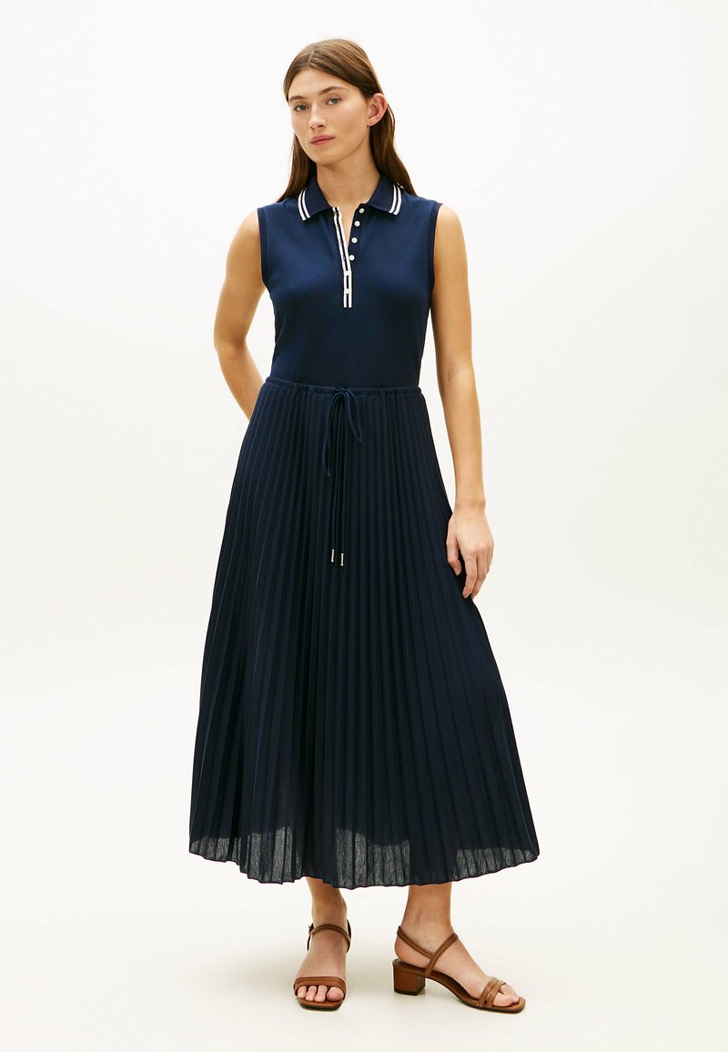 Femme portant une robe polo bleu marine sans manches avec une jupe plissée et des sandales à talons blocs ouvertes marron, debout devant un fond uni.