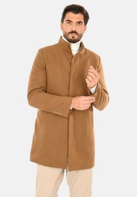 Cappotto in misto lana marrone con colletto alto, chiusura con bottoni, due tasche laterali e tessuto strutturato, indossato sopra un dolcevita bianco.