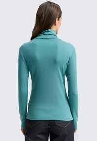 Langarmshirt in Teal mit hohem Kragen, glatter Textur und tailliertem Design, kombiniert mit dunklen Jeans. Keine sichtbaren Muster oder Akzente.