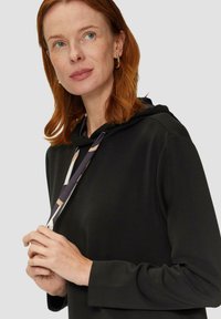 Zwarte hoodie van zacht materiaal, met een trekkoord in een geometrisch patroon in gedempte kleuren. Lange mouwen en een relaxte pasvorm.