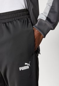 Person iført sorte Puma træningsbukser med elastisk talje og grå jakke, hånd placeret i bukselommen.