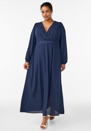 Naine kannab pikka tumesinist seotavat kleiti pikkade varrukatega ja beeže kõrgete kontsadega sandaale, seistes valgel taustal.