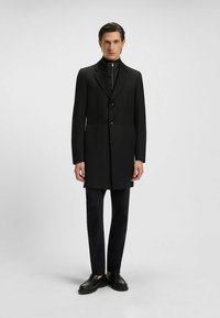 Cappotto nero con colletto a punta, chiusura con bottoni a un solo petto, tasche laterali, indossato sopra un top nero a collo alto e pantaloni slim.