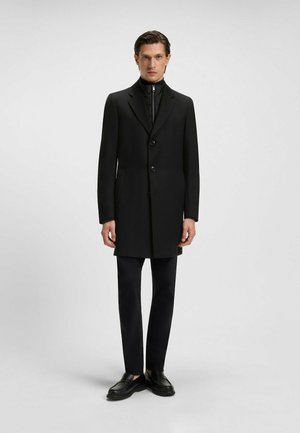 Black overcoat with a notched collar, single-breasted button closure, side pockets, worn over a black high-neck top and slim pants.