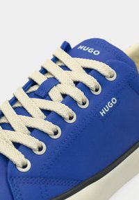 Μπλε συνθετικά αθλητικά παπούτσια με κρέμ κορδόνια. Διαθέτουν ασημένια ρέλια και λογότυπο HUGO. Η σόλα είναι μαύρη με υφή τελειώματος.