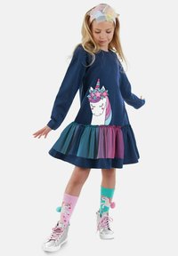 Mörkblå långärmad klänning med enhörningsgrafik, lager av tullekjol i teal och rosa, silverfärgade sneakers och färgglada strumpor med enhörningsdesign.