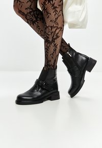 Bottines en cuir noir avec fermeture éclair sur le côté, talon épais et sangle décorative. Collants en résille texturée avec motif floral visible.