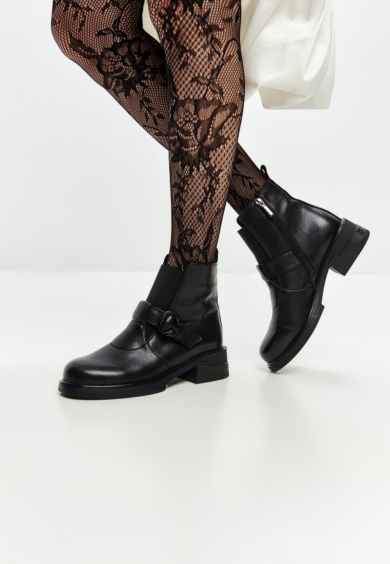 Bottines en cuir noir avec fermeture éclair sur le côté, talon épais et sangle décorative. Collants en résille texturée avec motif floral visible.