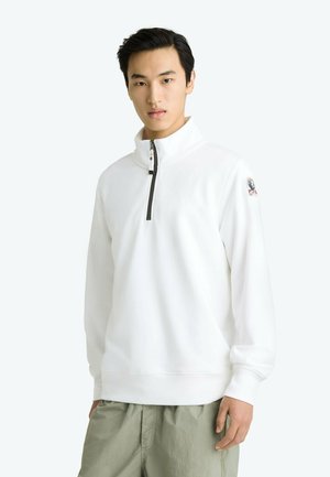 Homme portant un pull blanc à fermeture éclair quart de zip avec un patch logo sur la manche et un pantalon vert clair sur fond uni.