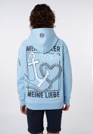 Person with curly hair wears light blue hoodie featuring large white anchor and heart-shaped rope design with German text on back.