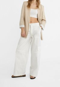 Beige blazer met opgerolde mouwen, witte crop top en losse witte cargobroek met zakken. Sandalen met getextureerde zolen maken de look compleet.