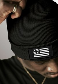 Black beanie with a ribbed texture and a rectangular tag featuring white stripes and two crosses, worn low on the forehead.
