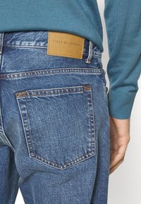 Los jeans de mezclilla en un lavado medio tienen una etiqueta de cuero marrón que dice "TIGER OF SWEDEN". Camiseta de punto azul de manga larga visible en el fondo.