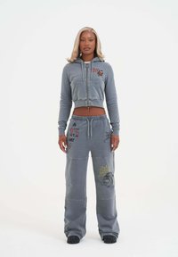 Kvinna som står vänd framåt i en grå kortärmad hoodie och matchande vida sweatpants med grafiska tryck och svarta skor.