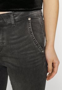 Primer plano de una persona que lleva puestos unos pantalones de mezclilla negros y una blusa negra, mostrando el bolsillo delantero y parte de la mano descansando al lado del muslo.