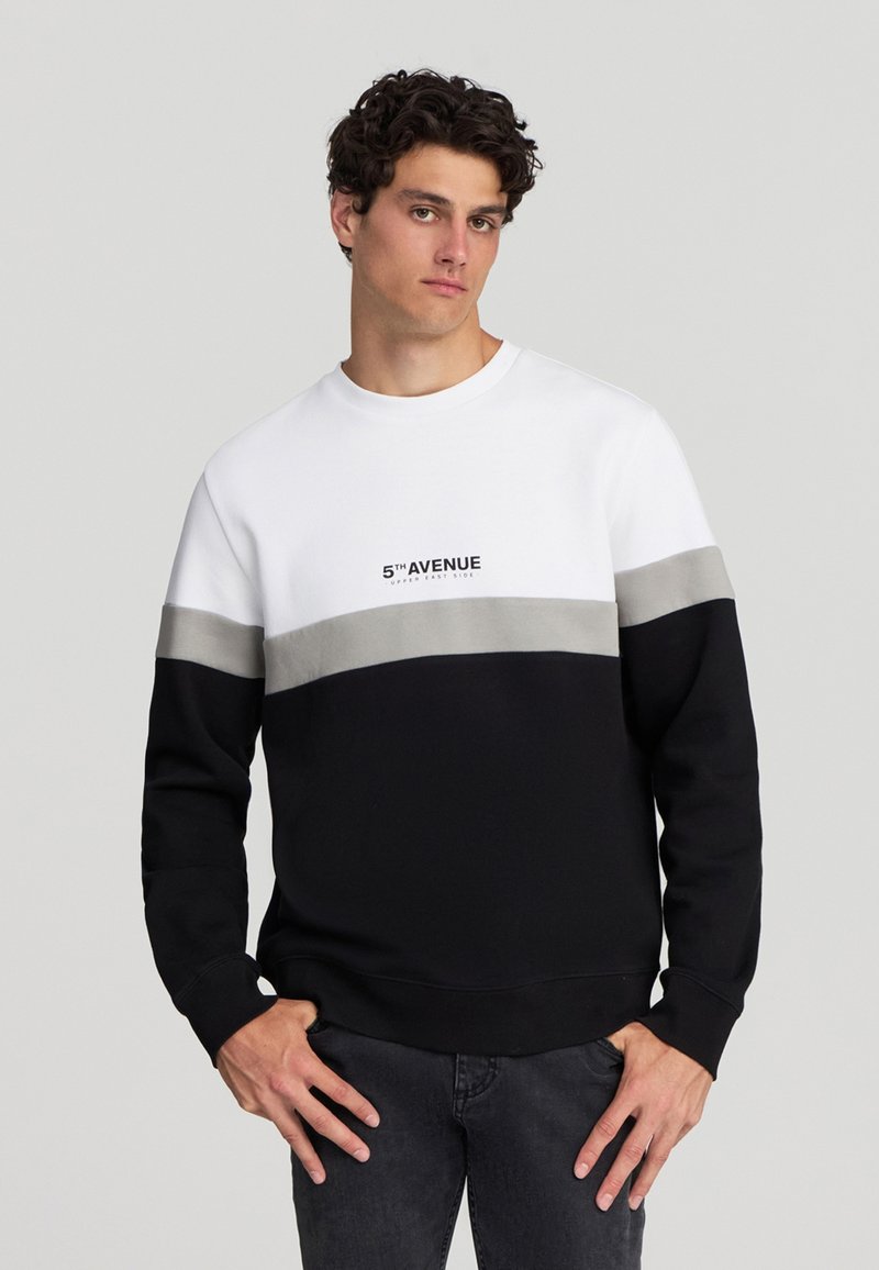 Giovane con una felpa a blocchi di colore bianca, grigia e nera con la scritta "5TH AVENUE", in piedi con le mani sui fianchi.