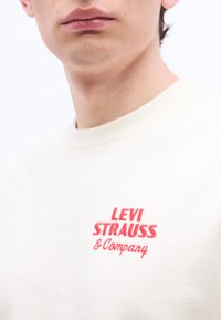 Κοντινό πλάνο ενός ατόμου που φοράει λευκό πουκάμισο με κόκκινο κείμενο «LEVI STRAUSS & Company» στο στήθος.