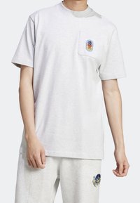 Osoba v světle šedém tričku s krátkým rukávem, na kterém je malá barevná nášivka na kapsičce, a odpovídajících světle šedých teplácích s emblémem.